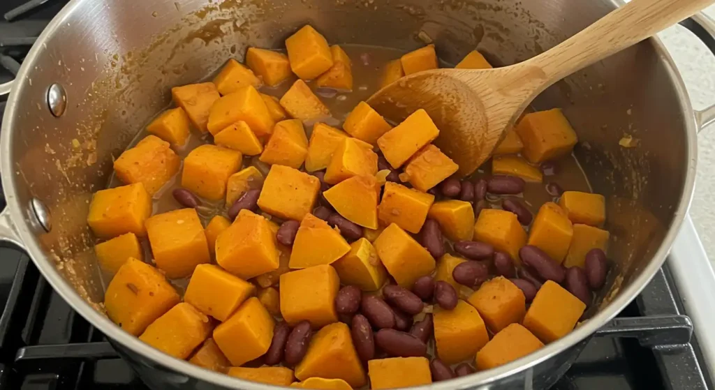 Kidney beans being added to the simmering pot of squash, with a swirl of coconut milk being stirred in for a creamy finish.