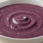 A vibrant bowl of purple black bean soup garnished with fresh cilantro and lime wedges, served in a rustic ceramic bowl.